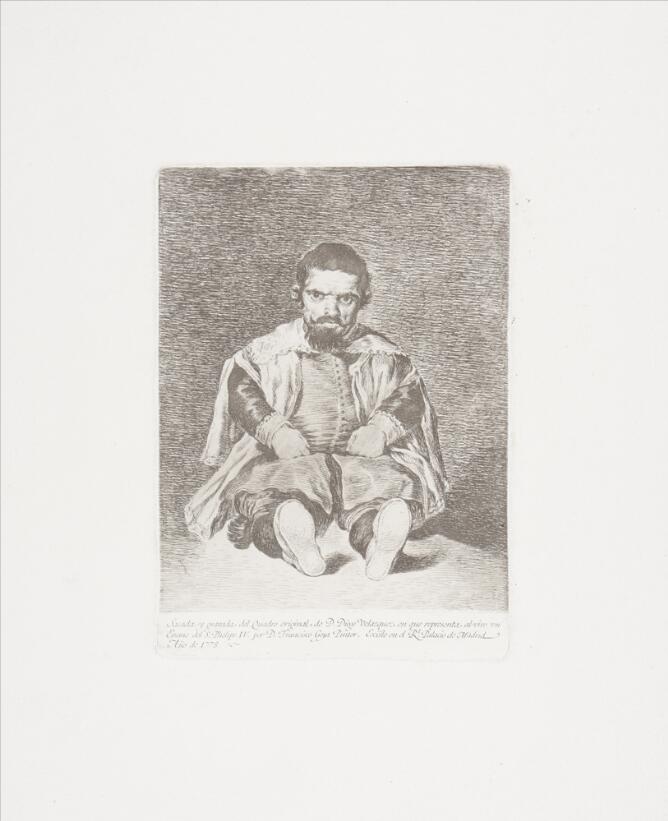 A black and white portrait of a man of short stature wearing a cloak, sitting with his legs in front of him and facing the viewer